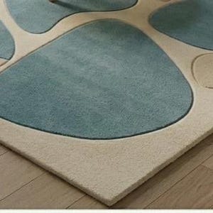 May include: A rectangular rug featuring a light beige border and a design of large, irregular shapes in shades of blue and teal. The rug is positioned on a wooden floor.
