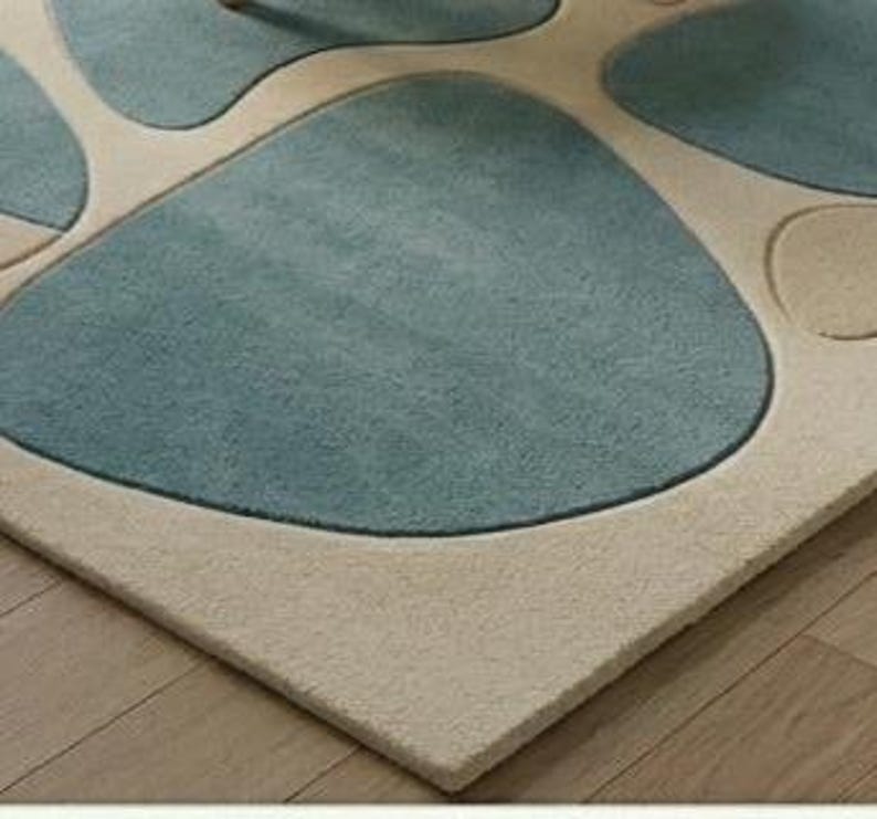 May include: A rectangular rug featuring a light beige border and a design of large, irregular shapes in shades of blue and teal. The rug is positioned on a wooden floor.