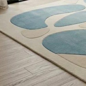 May include: A rectangular rug featuring a cream backdrop with abstract blue and beige shapes. The rug rests on a light-coloured wooden floor, with a glimpse of a white curtain in the top left corner.