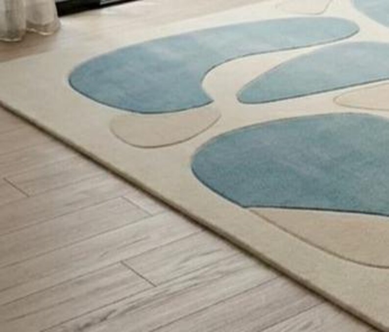May include: A rectangular rug featuring a cream backdrop with abstract blue and beige shapes. The rug rests on a light-coloured wooden floor, with a glimpse of a white curtain in the top left corner.