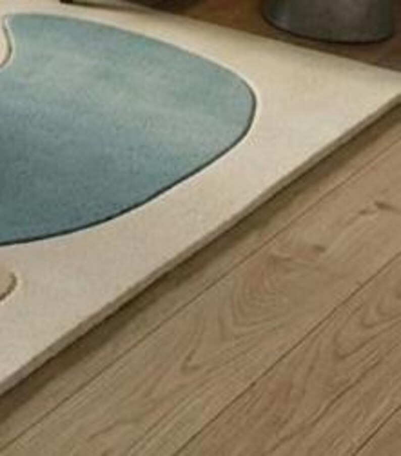 May include: A close-up of a rug featuring a light blue, kidney-shaped design on a cream background, with a dark blue outline. The rug is placed on a wooden floor.