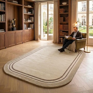 May include: A large, rectangular area rug with rounded corners, featuring a solid beige center and a double-line border in darker brown tones. The rug is placed on a light-colored wooden floor in a room with built-in wooden bookshelves.