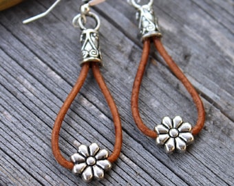 May include: A pair of brown leather earrings with silver flower charms and silver hooks. The earrings are hanging from a wooden surface.