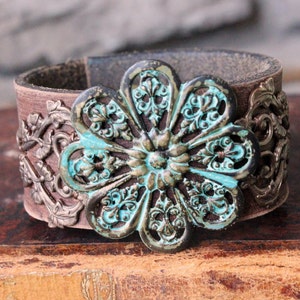 May include: A brown leather cuff bracelet with a large, ornate metal flower in the center. The flower is turquoise and gold with a distressed finish.