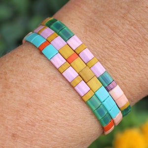 May include: Three colourful beaded bracelets in shades of turquoise, green, pink, orange, and gold. The bracelets are made of small, rectangular beads and are stacked on a wrist. The background is blurred, with hints of green and yellow.