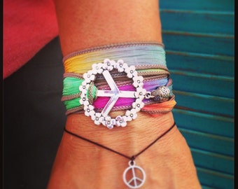 Könnte beinhalten: Ein mehrlagiges Armband mit einem leuchtend regenbogenfarbenen Stoffband, das um das Handgelenk gewickelt ist, verziert mit einem großen silberfarbenen Friedenszeichen-Anhänger, der von kleinen Blumenmotiven umgeben ist, sowie einem dünnen schwarzen Kordelarmband mit einem kleinen Friedenszeichen.
