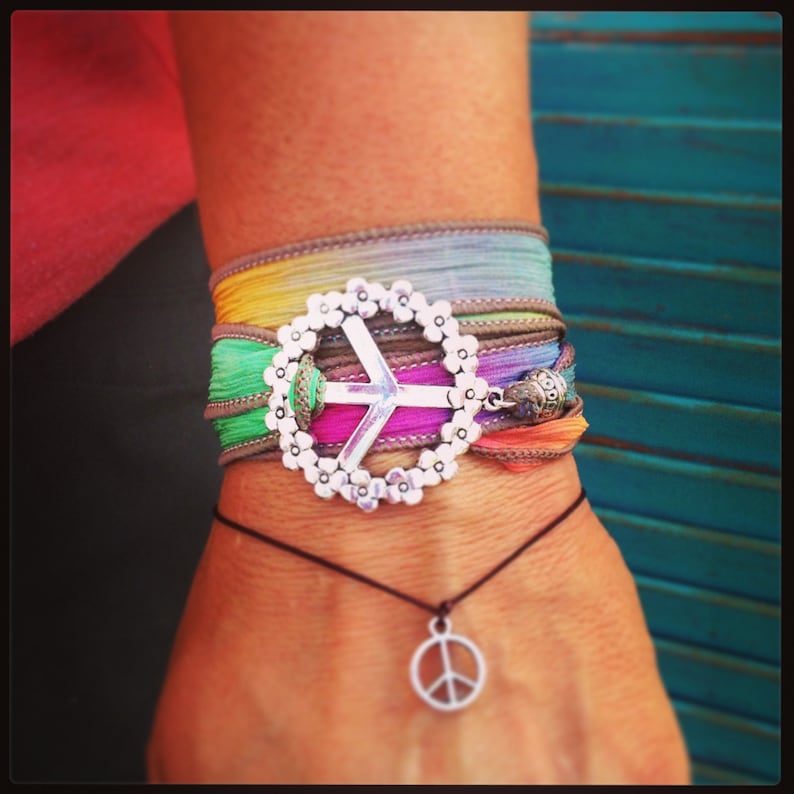 May include: A multi-colored wrap bracelet with a silver peace sign charm and a matching peace sign pendant necklace. The bracelet features a floral design around the peace sign and is made of colorful ribbons.