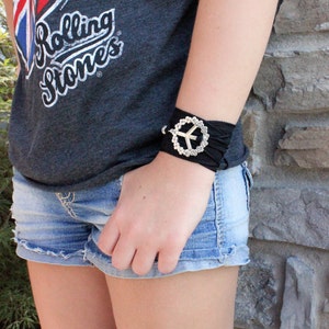 May include: A black fabric bracelet with a silver peace sign charm. The bracelet is worn on a person's wrist. The person is wearing a gray t-shirt with "Rolling Stones" printed on it and denim shorts.