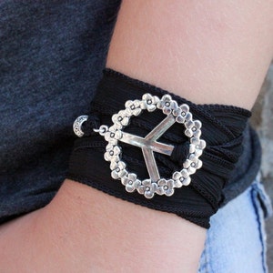 May include: Black wrap bracelet featuring a silver peace sign charm. The peace sign is surrounded by a floral design. The bracelet is wrapped around the wrist and secured with a small silver clasp.