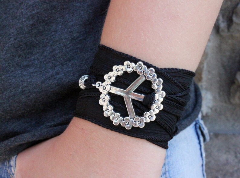 May include: Black wrap bracelet featuring a silver peace sign charm. The peace sign is surrounded by a floral design. The bracelet is wrapped around the wrist and secured with a small silver clasp.