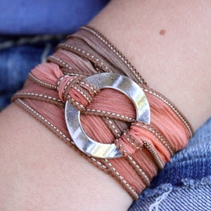 May include: A wrap bracelet featuring peach and brown fabric ribbons with white stitching. The bracelet is secured with a silver-colored circular clasp. The accessory is wrapped around the wrist.