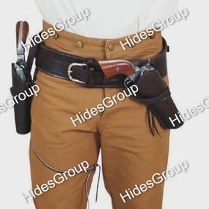 Könnte beinhalten: Ein dunkelbrauner Leder-Waffengürtel mit zwei passenden Holstern, die jeweils einen Revolver mit Holzgriff enthalten. Der Gürtel hat eine silberfarbene Metallschnalle und wird über einer beigen Hose und einem weißen Hemd getragen. Der Text HidesGroup ist sichtbar.