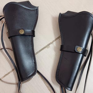 Könnte beinhalten: Zwei schwarze Leder-Pistolenholster mit dekorativen messingfarbenen Knöpfen und Riemen. Die Holster sind für das Tragen an der Hüfte konzipiert und haben einen klassischen Western-Stil. Die Holster sind aus strapazierfähigem Leder gefertigt.