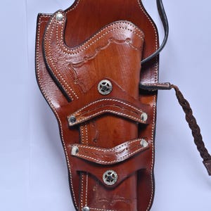 Puede incluir: Una funda de cuero marrón con costuras decorativas y detalles plateados en forma de estrella. La funda tiene un lazo para un cinturón y una correa de cuero trenzado. La funda está diseñada para guardar un arma de fuego.