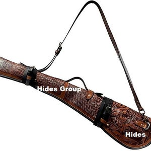 以下が含まれることがあります： 茶色の革製ライフルケース。装飾的な花の模様と「Hides Group」と「Hides」の文字が入っています。ケースには黒いショルダーストラップとバックル留めが付いています。このケースは長銃を運ぶように設計されています。