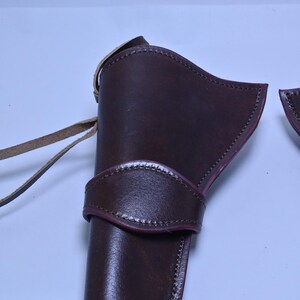 May include: A dark brown leather holster with a strap and belt loop. The holster has a curved design and is stitched along the edges. The leather has a glossy finish. The holster is designed to hold a firearm.