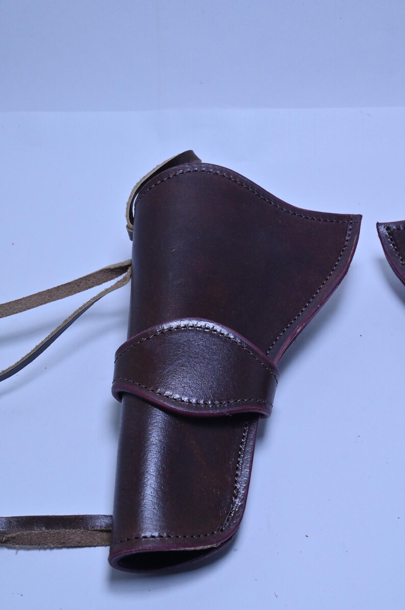 May include: A dark brown leather holster with a strap and belt loop. The holster has a curved design and is stitched along the edges. The leather has a glossy finish. The holster is designed to hold a firearm.