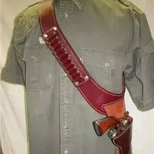 May include: A burgundy leather shoulder holster with a matching ammunition belt. The holster is designed to carry a revolver. The belt has multiple loops for holding bullets. The holster is worn over a short-sleeved olive green shirt.