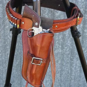 May include: A brown leather holster and matching belt featuring a series of cartridge loops holding metallic ammunition. The holster contains a silver-toned revolver with a dark wood grip, secured by a leather strap with a metal buckle.