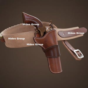 Op de afbeelding: Een bruin leren holster- en riemset met een revolver. De holster is bevestigd aan een beige riem met een bruin lederen accent. De revolver heeft een houten handvat. De tekst "Hides Group" is zichtbaar.