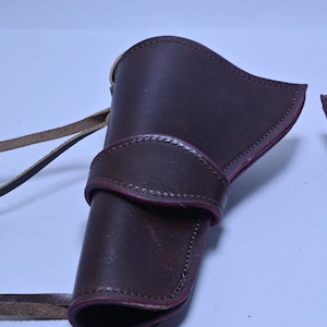 May include: A dark brown leather holster with a curved top and a strap. The holster has a stitched design and a belt loop. The leather is a rich, deep brown colour.