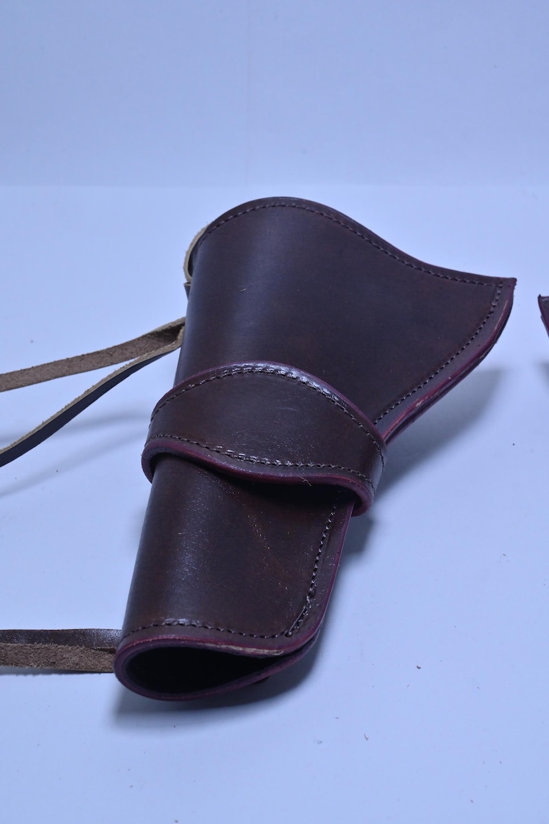 May include: A dark brown leather holster with a curved top and a strap. The holster has a stitched design and a belt loop. The leather is a rich, deep brown colour.