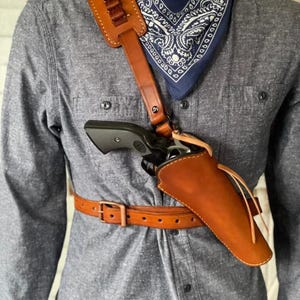 Könnte beinhalten: Ein braunes Leder-Schulterholster mit passendem Gürtel. Das Holster hält einen schwarzen Revolver. Der Schulterriemen hat Schlaufen für Patronen. Ein blaues Paisley-Bandana ist sichtbar.