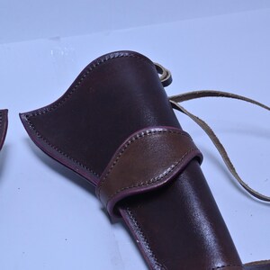 May include: A brown leather holster with a contrasting brown strap and stitching. The holster has a curved design and a loop for attaching to a belt. The leather has a glossy finish.