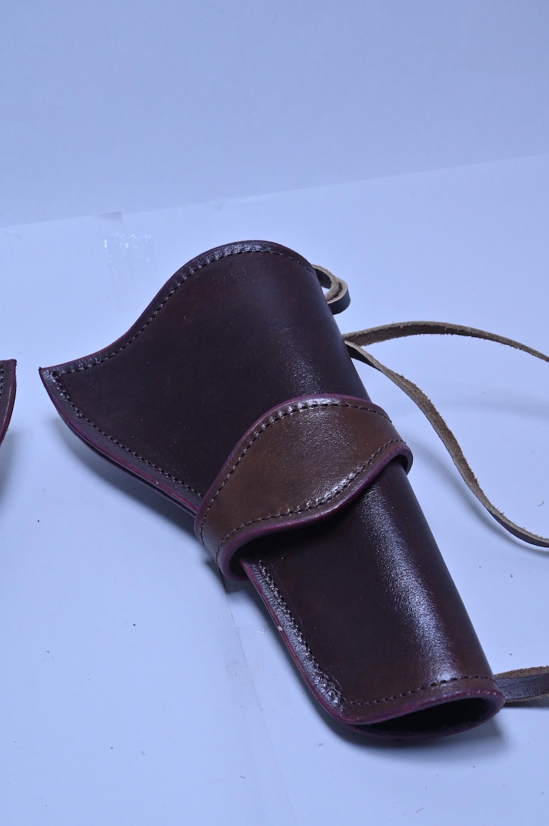 May include: A brown leather holster with a contrasting brown strap and stitching. The holster has a curved design and a loop for attaching to a belt. The leather has a glossy finish.