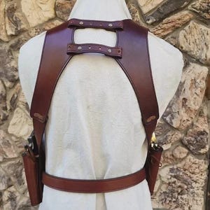 Op de afbeelding: Een donkerbruin lederen schouderholster-tuig met twee zijholsters, een heupriem en een rugsteun verbonden door twee horizontale banden met metalen klinknagels. Het holstersysteem wordt over een wit kledingstuk gedragen en heeft gestikte randen.