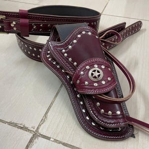 Puede incluir: Juego de funda de pistola y cinturón de cuero burdeos. La funda presenta un emblema de estrella decorativo y tachuelas plateadas. El cinturón tiene una hebilla y detalles de tachuelas a juego. El conjunto está diseñado para un look de temática occidental.
