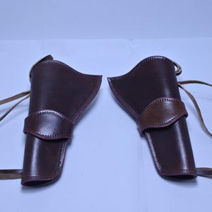 May include: Two brown leather gun holsters with tan straps. The holsters are a deep reddish-brown colour with a lighter brown accent on the belt loop. The straps are long and thin.