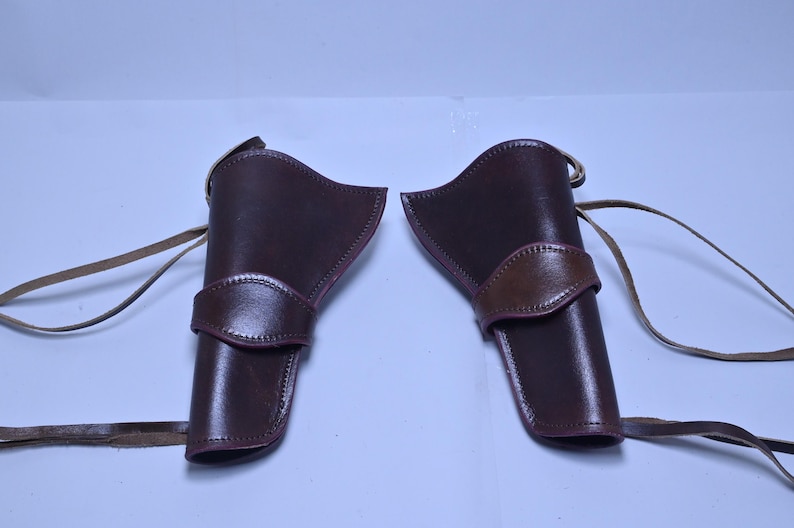 May include: Two brown leather gun holsters with tan straps. The holsters are a deep reddish-brown colour with a lighter brown accent on the belt loop. The straps are long and thin.