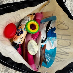 Może przedstawiać: Płócienna torba typu tote z czarnym zamkiem błyskawicznym i wykończeniem, wypełniona różnymi przedmiotami. Zawartość: czerwona pokrywka, tubka kremu do rąk, zielono-żółty pierścień, różowy przedmiot i niebieska torebka.