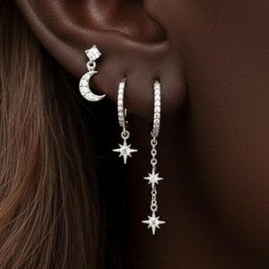 May include: A collection of silver earrings featuring celestial designs. One earring has a small diamond-shaped stud, another has a crescent moon, and two hoop earrings with star pendants. The earrings are set against a dark background.