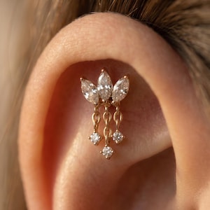 Può includere: Un orecchino dorato con tre gemme a forma di marquise nella parte superiore e tre gemme rotonde pendenti. L'orecchino è attaccato all'orecchio. Le gemme sono chiare e scintillanti.