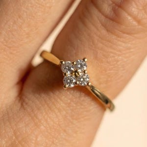 Könnte beinhalten: Ein goldener Ring mit einem vierblättrigen Kleeblattmuster aus kleinen, runden Diamanten. Der Ring hat ein dünnes, poliertes Goldband. Die Diamanten sind eng beieinander gesetzt und erzeugen einen funkelnden Effekt auf dem Gold.