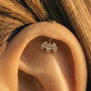 May include: Close-up of an ear featuring a gold helix piercing with a cluster of three marquise-cut diamonds. The jewellery is set against the skin and hair, with the diamonds sparkling.