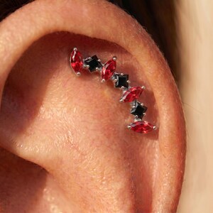 Könnte beinhalten: Ein silberfarbenes Knorpel-Piercing für das Ohr mit einem abwechselnden Muster aus drei roten Edelsteinen im Marquise-Schliff und drei schwarzen Edelsteinen im quadratischen Schliff, angeordnet in einer geschwungenen Linie.