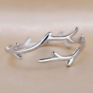 May include: A silver-colored ring with a branch-like design. The ring features multiple curved lines that resemble twigs or branches, creating an organic and natural aesthetic. The ring is set against a neutral background.