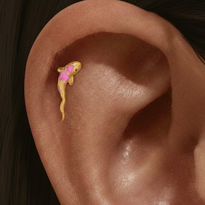 May include: A gold-coloured fish-shaped earring with pink detailing. The earring is a small, detailed design, suitable for a cartilage piercing. The fish has a curved body and a long, flowing tail.