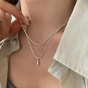 May include: A delicate silver necklace with two strands. The top strand has a beaded design, while the bottom strand features a teardrop pendant. The necklace is worn against a neutral background, highlighting its simple elegance.