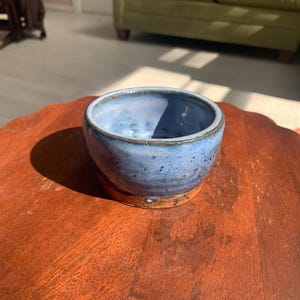 May include: A small, round ceramic bowl featuring a speckled light blue glaze on the upper portion and a smooth, unglazed terracotta-colored base. The interior of the bowl shows a matching light blue finish with subtle variations in color.