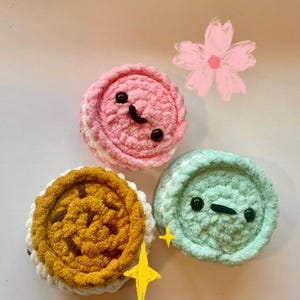May include: Three crocheted macarons in pink, yellow, and mint green, each with a white filling and a smiling face. The pink macaron has a curved smile, while the mint green one has a straight line. A yellow star and a pink flower are also in the image.