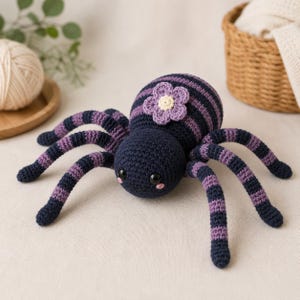 Peut inclure: Un jouet araignée en crochet avec un corps rond rayé de bleu marine foncé et de violet. L'araignée possède huit pattes rayées, deux yeux en plastique noir, de petites marques de blush rose et une fleur violet clair au centre crème.