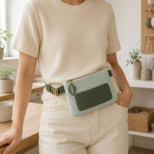 Peut inclure: Un sac banane en crochet vert clair avec une poche frontale rectangulaire vert foncé, une fermeture éclair dorée et une petite breloque en forme de feuille. Le sac est fixé à une sangle réglable vert foncé avec une boucle dorée.