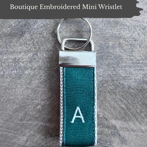 May include: A teal mini wristlet with a silver key ring and clasp. The wristlet is embroidered with a white letter "A". The text "Boutique Embroidered Mini Wristlet" is at the top. The text "Custom Made • Boutique Quality Durable & Stylish" is at the bottom.