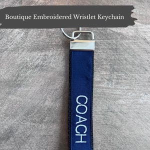 May include: A navy blue wristlet keychain with the word "COACH" embroidered in white. The keychain has a silver-coloured metal clasp. The text "Boutique Embroidered Wristlet Keychain" is at the top of the image.