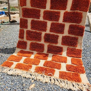May include: A rectangular, hand-woven rug with a brick-like pattern in shades of orange and cream. The rug features a textured, tufted design and a fringed edge. This decorative rug is suitable for home decor.