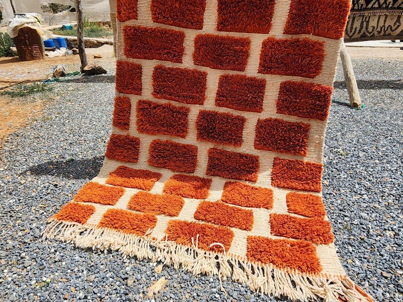May include: A rectangular, hand-woven rug with a brick-like pattern in shades of orange and cream. The rug features a textured, tufted design and a fringed edge. This decorative rug is suitable for home decor.